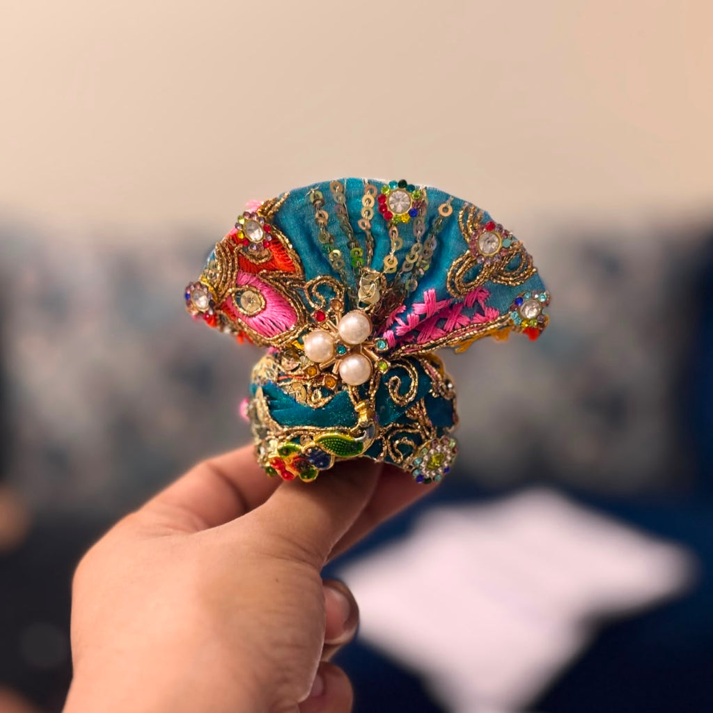Handcrafted vibrant teal green designer dress and matching crown for Ladoo Gopal Ji, featuring multi-colored thread embroidery and pearl borders.