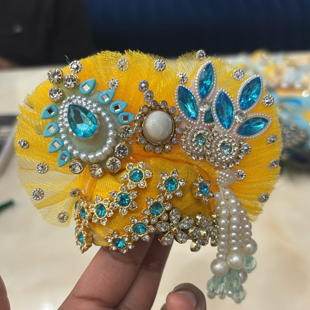 Handcrafted peacock-themed designer dress and matching crown for Ladoo Gopal Ji, featuring blue and yellow petal layers and teal crystal accents.