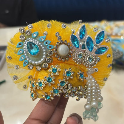 Handcrafted peacock-themed designer dress and matching crown for Ladoo Gopal Ji, featuring blue and yellow petal layers and teal crystal accents.