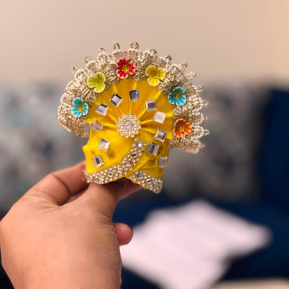Handcrafted fuchsia pink designer dress and matching yellow crown for Ladoo Gopal Ji, featuring peacock-plume borders and blue gemstone accents.