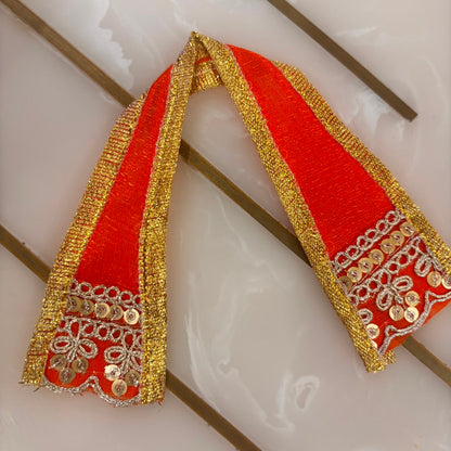 A bright orange fabric ribbon or sash with ornate gold metallic trim and mirror work, laid flat on a glossy white surface.