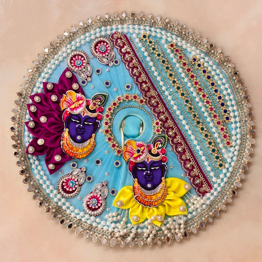 Handcrafted sky-blue designer dress and matching crown for Ladoo Gopal Ji, featuring Radha-Krishna figures, purple petal folds, and pearl detailing.